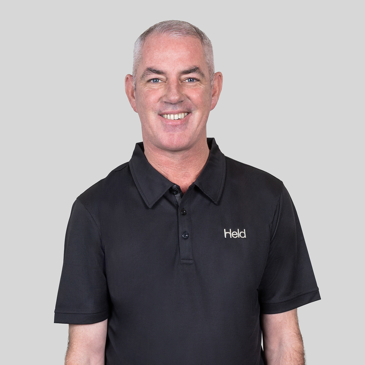 Smiling man in a black polo shirt with a small Held logo, standing against a light gray background.
