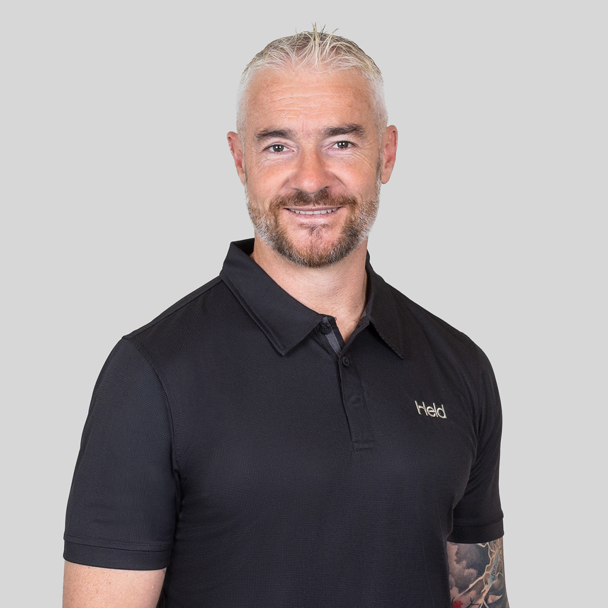 Portrait of a smiling man with short gray hair and a beard, wearing a black polo shirt against a light gray background.