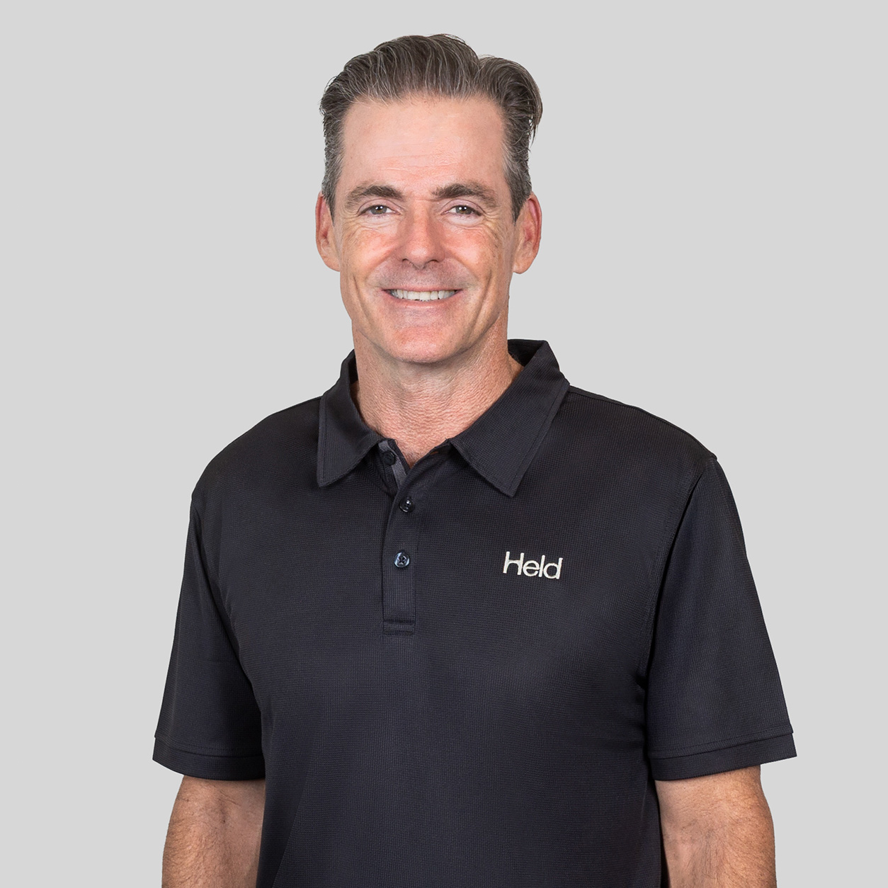 Smiling man in a black polo shirt with a small Held logo, posed against a gray background.