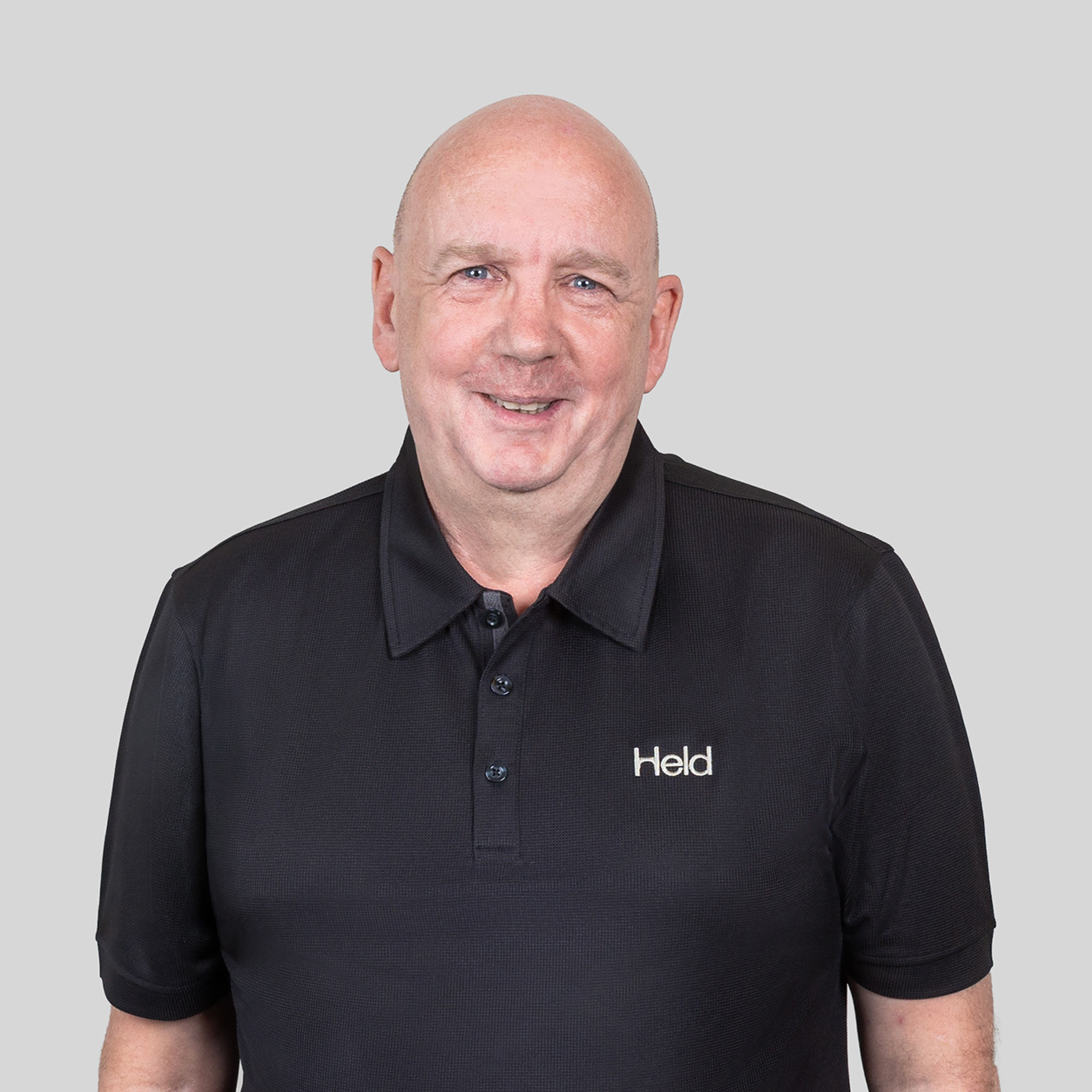 Smiling bald man wearing a black polo shirt with 'Held' logo, neutral gray background.